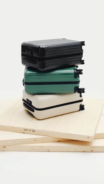 The image features a stylized product shot of three stacked mini-suitcases in different colors: black, green, and cream. They are arranged on top of two light-colored wooden boards, with the boards at an angle to each other and in the lower portion of the image. The suitcases are a glossy material, and have black accents and small black wheels. The background is a clean white.