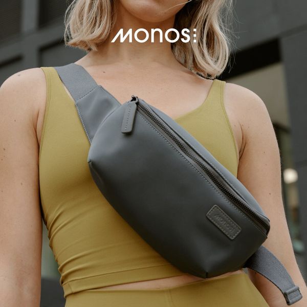 A woman is modeling a grey waist bag. She is wearing a yellow tank top and matching tights against an urban background. The waist bag is across her chest, and appears to be made of a smooth material. The brand logo is displayed above the model.
