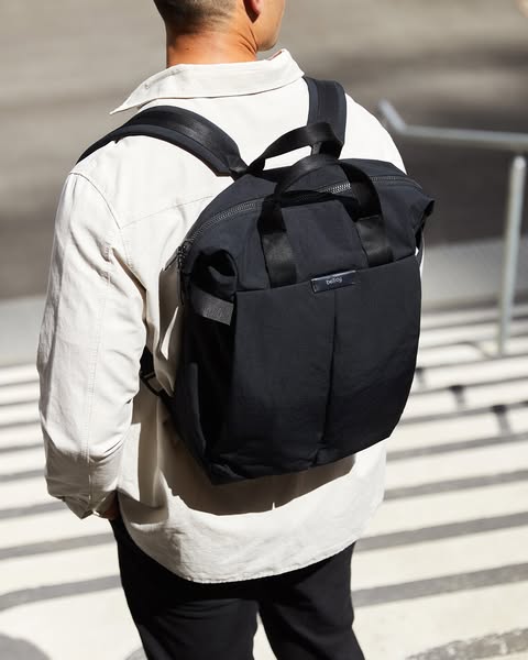 A person wearing a light-colored shirt and black pants is seen from behind, walking on an outdoor structure comprised of steps. They are wearing a black backpack/tote hybrid bag over their shoulders. The background is blurred, with glimpses of neutral-toned buildings and sunlight.