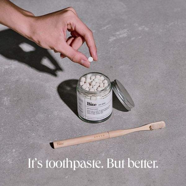 The image showcases a minimalist product presentation. A hand is holding a single, small white tablet above a glass jar filled with similar tablets. The jar has a silver lid and is labeled 'Bite 245 BITES' with instructions and 'NATURALLY WHITENING TOOTHPASTE'. Below the jar is a wooden toothbrush. The entire scene is set against a neutral, gray background. The ad copy 'It's toothpaste. But better.' is displayed at the bottom.
