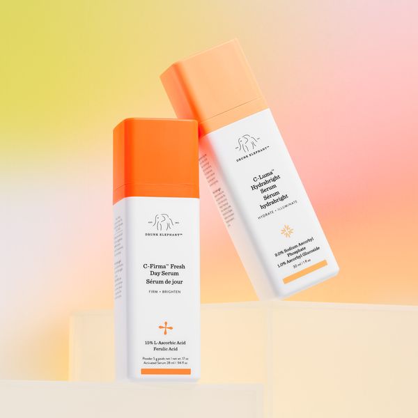 The image showcases two serum bottles against a gradient background of yellow, orange, and pink. The bottles are white with orange caps and labels. One bottle is upright, while the other is tilted, creating a dynamic composition. The text on the bottles highlights key ingredients and product benefits. The overall aesthetic is clean, modern, and product-focused, with attention to skincare and beauty.