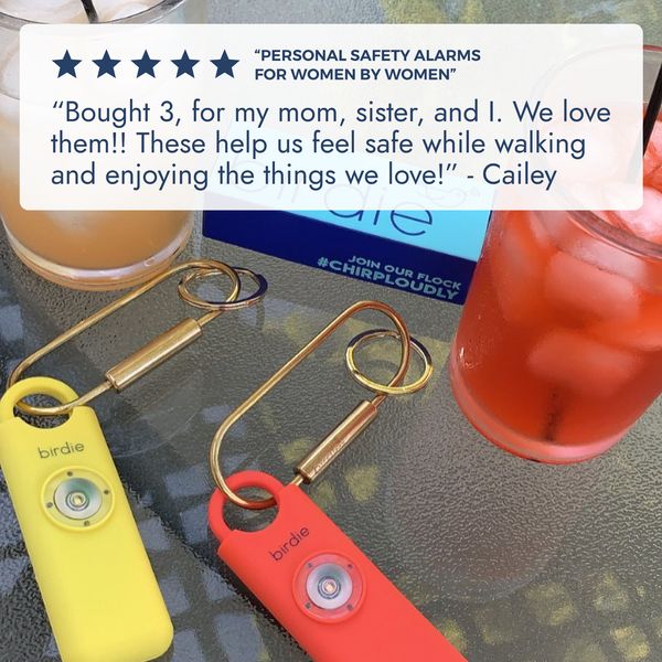 The ad is a promotional image showcasing personal safety alarms. Two alarms are displayed, one yellow and one red, resting on a glass table. A testimonial in a speech bubble, with star ratings, provides customer feedback, conveying the alarm's effectiveness in helping individuals feel safe during activities. Drinks and keychains in the background suggest a casual scene.