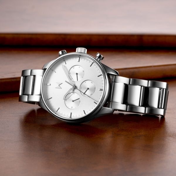 A close-up shot of a silver wristwatch with a white face, resting on a polished wooden surface. The watch has a stainless steel band and chronograph subdials. The image is well-lit, emphasizing the watch's details and the reflective quality of the metal. The layout is clean and focused on the watch as the sole subject, with a subtle, blurred background.