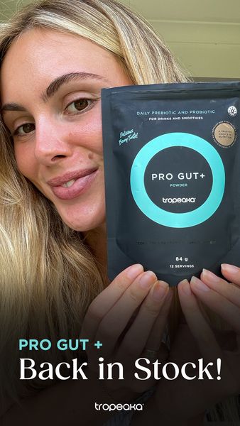 An ad featuring a fair-skinned blonde woman smiling and holding a black bag of 'PRO GUT+' powder. The woman is centered and the bag is in front of her. The bag has turquoise circular graphics and text. The background is a neutral wall.  The text at the bottom reads, 'PRO GUT + Back in Stock! '. The ad's overall tone is bright, positive, and inviting.