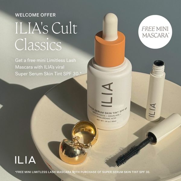 The ad features a product flat lay on a cream-colored surface. The primary focus is on two white bottles of skincare products, one with a dropper and the other a mascara. The mascara has a visible wand. Above the products is white text that reads "WELCOME OFFER" and "ILIA's Cult Classics". There is additional text that says "Get a free mini Limitless Lash/ Mascara with ILIA's viral / Super Serum Skin Tint SPF 30.*". A small gold ring sits in front of the bottles. The background is a soft, neutral tone to highlight the products. The layout is clean and minimal, emphasizing the products and the offer.