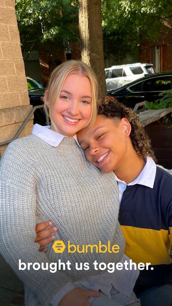 An image featuring two young women embracing and smiling at the camera. One woman, with blonde hair, is wearing a gray sweater and white collared shirt, embracing the other, who is wearing a navy blue, yellow, and white striped rugby shirt. The background suggests an outdoor setting.