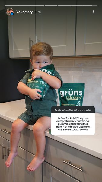 An ad featuring a young child sitting on a countertop holding a bag of "grüns kids daily" gummies. The child is wearing a matching green outfit. To the right of the child are two bags of the product. A text box provides information on the product, stating it helps kids eat more veggies and that the child in the image loves them.