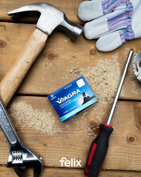 An overhead shot shows a package of "Viagra" surrounded by construction tools such as a hammer, adjustable wrench, gloves, a screwdriver, and sawing dust strewn across a wooden plank background. The package is in a blue box with the text "VIAGRA" in white at the top, and a silhouette of a person on a mountain in the side, and the text "50 mg" shows at the bottom. The layout indicates a comparison implying strength or construction capabilities connected in a metaphoric way.
