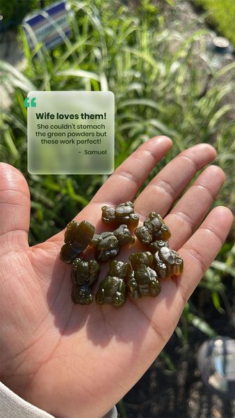 A person's hand is holding a handful of green, gummy bear-shaped supplements. A semi-transparent square with a quote is superimposed on the upper left with a quote from Samuel that the person's wife loves them (referring to the supplements) because she couldn't stomach the green powders. The background is slightly blurred with green foliage.