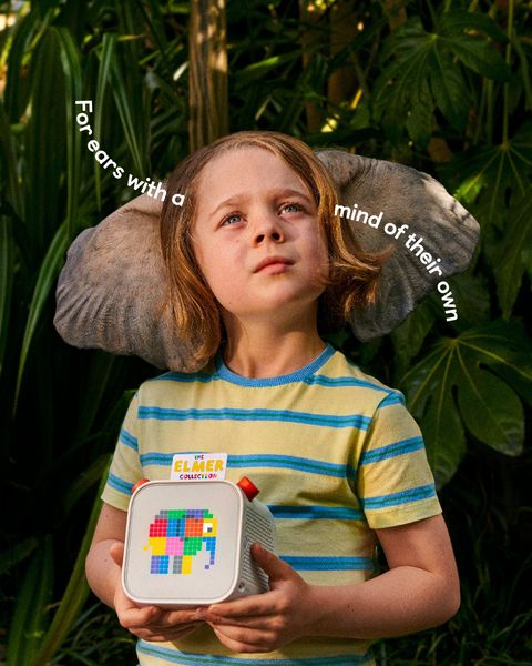 A young child is the central figure with the head partially obscured by large elephant ears. The child is looking upwards with a thoughtful expression, set against a lush green background with large leaves. In their hands, the child holds a small, colorful device. The text on the device, "The Elmer Collection",  suggests a product designed with the child in mind. The overall composition evokes a sense of childhood wonder and imagination, enhanced by the text "For ears with a mind of their own".