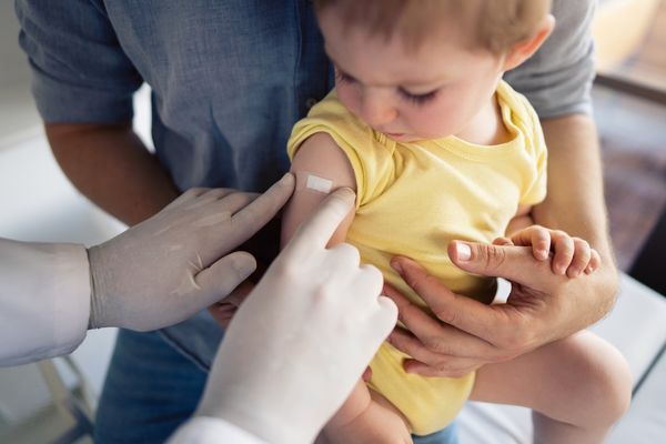 A photograph shows a baby in a yellow onesie being held by an adult, likely a parent. The baby's arm has a bandage on it, suggesting a recent vaccination. A medical professional, wearing a white coat and gloves, is pointing to the bandage, while another adult's hand is gently holding the baby's free arm. The overall composition suggests a comforting and reassuring scene.
