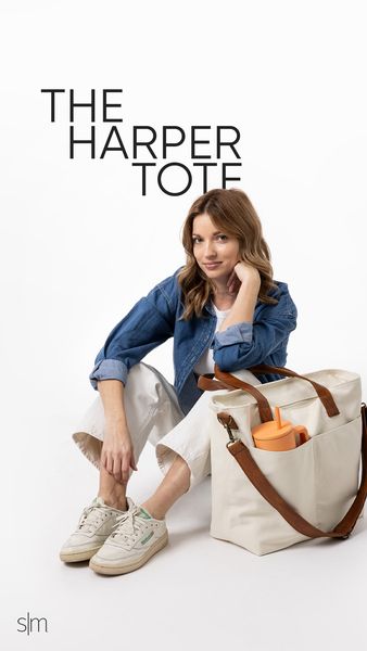 A lifestyle shot featuring a woman in casual clothes sitting on a white background. She is wearing a denim jacket, white pants, and sneakers. Next to her is a large tote bag with brown leather straps. The tote bag has an outside pocket and contains a child's orange sippy cup. The ad's layout includes simple text at the top that reads "THE HARPER TOTE". The background is a plain white.