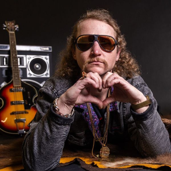 A studio shot featuring a man in a vintage style. He wears sunglasses, a denim jacket, and gold jewelry including multiple necklaces. He forms a heart shape with his hands. In the background, there is a bass guitar, a boombox, and a taxidermy-style surface, contributing to the retro aesthetic.