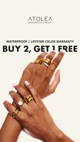 An advertisement featuring two hands adorned with gold rings on a cream background. The hands are positioned to showcase various ring designs. The text at the top includes brand name and product information, followed by the promotional offer in large, bold letters: BUY 2, GET 1 FREE. Each hand displays multiple rings, showcasing different styles. The overall composition is clean with a focus on the jewelry. The lighting is bright, highlighting the gold color of the rings.