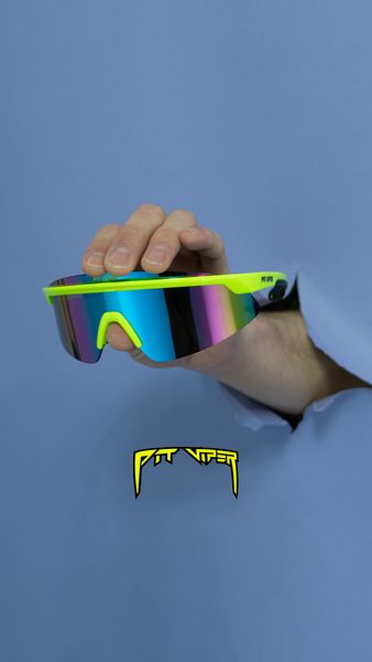 An eye-level shot shows a pair of sunglasses being held by a hand that is partially ripped through a blue paper background. The sunglasses have neon green frames and mirrored lenses with blue to pink gradient. The brand name is visible. The background is a solid light blue color. Below the sunglasses is another brand writing in yellow.