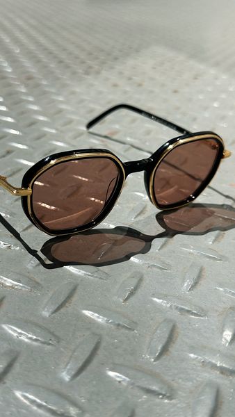 A close-up shot of a pair of sunglasses resting on a textured, metallic surface. The sunglasses have a sleek black frame with golden accents around the lenses and temples. The lenses themselves are tinted in a shade of brown. The background is a well-lit, industrial-looking diamond-plate metal surface. The glasses cast a crisp shadow, enhancing their presence and design.