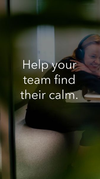 The image is a vertically oriented advertisement. It features a young woman with long, reddish-blonde hair wearing headphones, leaning on a desk with a relaxed posture and a gentle smile. The background is a soft mix of muted greens and grays, possibly the interior of an office. The text overlayed on the image reads, 'Help your team find their calm.' There are blurred foreground elements on both sides of the image adding depth and focus to the center. The composition is intended to promote a product or service that facilitates workplace wellness.