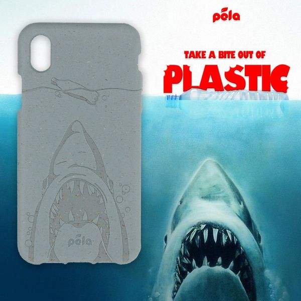 The ad features a smartphone case with a shark design, juxtaposed with a separate image of a shark's open mouth. Above the shark case and on the right side of the image is the text "TAKE A BITE OUT OF PLASTIC" in a red, bold font. The phone case has an engraved design displaying both a shark and a plastic bottle. The case is slightly angled, suggesting a 3D representation. The background continues the underwater theme with blue water. The sharks mouth is open as if to 'bite' at the viewer.