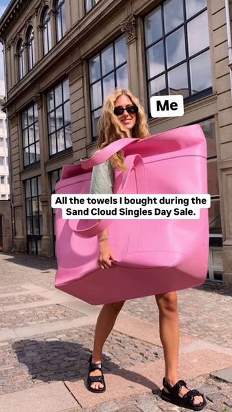 A woman with blonde hair and sunglasses is smiling and carrying an oversized pink bag. The bag is so large that it almost completely obscures her body. Text overlayed on the image says 'Me' and 'All the towels I bought during the Sand Cloud Singles Day Sale.' She is standing in front of a building with large windows.