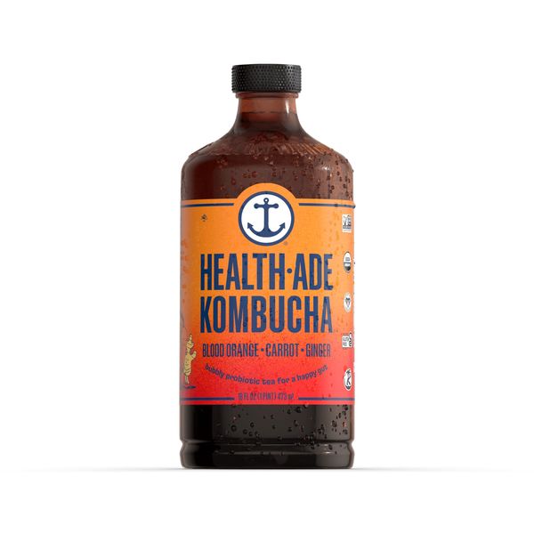 A close-up studio shot of a bottle of kombucha. The bottle is made of dark glass and filled with a dark, reddish-brown liquid. The label is orange with white and dark blue text, and an anchor logo. The text reads 'HEALTH-ADE KOMBUCHA' with another line of text below that reads 'BLOOD ORANGE - CARROT - GINGER, bubbly probiotic tea for a happy gut'. The bottle is partially covered in condensation, suggesting it's cold. The background is plain white, isolating the bottle and highlighting the product.