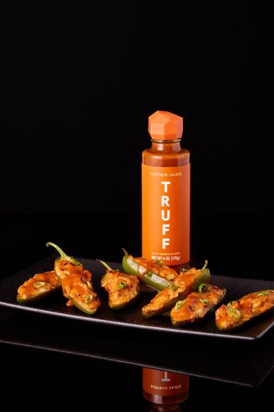 The image showcases a bottle of buffalo sauce with the brand name prominently displayed, alongside a black rectangular plate holding several jalapeno poppers. The poppers are filled and topped with sauce. The background is a stark black, accentuating the vibrant colors of the sauce and the food.