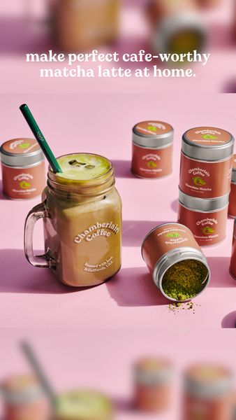 A product shot on a pink background featuring a matcha latte in a mason jar with a green straw, surrounded by several small cylindrical containers of matcha powder. One container is open, spilling the powder. The image is well-lit and has a clean, minimalist style. The top of the image contains text promoting the matcha latte.