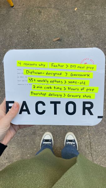 The image shows a person holding a white package with the word "FACTOR" printed in large, bold, black letters at the bottom. The package has handwritten content highlighted with yellow markers. At the top are the words "4 reasons why" which lead to a list comparing "Factor" to other ways of meal prepping. The comparisons are clearly written and include: "Factor > DIY meal prep", "Dietician-designed > Guesswork", "35+ weekly options > same-old", "2 min cook time > Hours of prep", and "Doorstep delivery > Grocery store". The viewer's feet and lower legs are visible suggesting the image was taken from a first-person perspective.