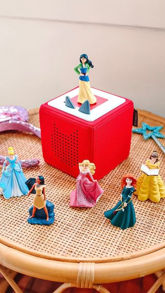 An image featuring a red cube-shaped audio player with several princess figurines displayed around it. Mulan stands atop the cube. Other princesses, including Cinderella, Pocahontas, Aurora, Merida, and Belle, are arranged nearby. A decorative star-shaped wand sits to the right of the cube. The scene is set on a tan wicker surface.