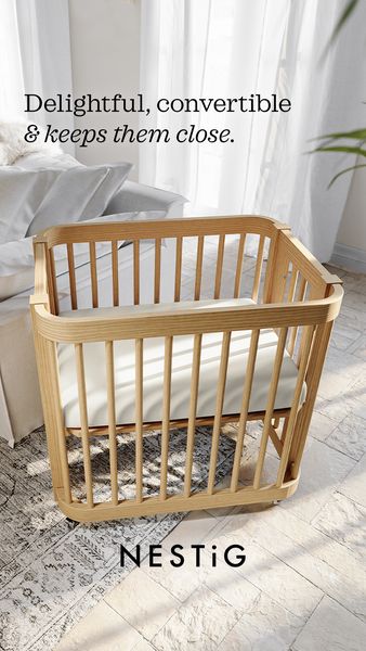 The advertisement features a light wooden crib set in a bright, airy room. The crib has a rectangular shape with vertical bars for sides and a simple, modern design. It's placed on a patterned rug, with a bed in the background and soft, white curtains. The overall aesthetic is clean and minimalist, and the angle is shot from above, showcasing the crib and its surroundings. The text overlay says 'Delightful, convertible & keeps them close.' at the top and brand name is on the bottom.