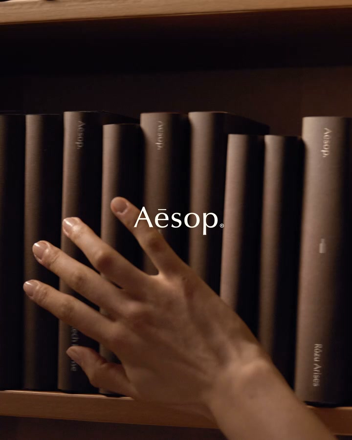 A close-up shot shows a person's hand gently touching a row of dark-colored books on a shelf. The Aesop logo is prominently displayed in white text over the books. The copy overlays the image, inviting viewers to explore a range of fragrances designed to evoke feelings and thoughts.