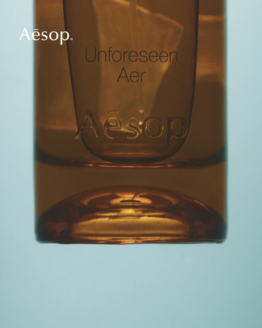 A close-up, amber-colored glass bottle of Aesop perfume is shown against a soft blue background. The bottle has the brand name 'Aesop' embossed on it, and the text 'Unforeseen Aer' is printed on the glass. The copy overlays the image, introducing the new fragrance as an aromatic fable about the value of imperfection and announcing its availability in stores and online.