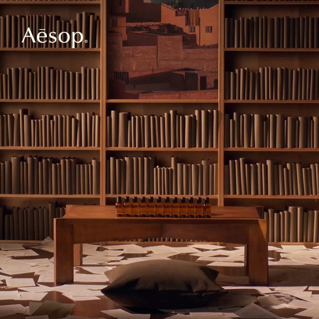 The image displays a warm, inviting scene with a bookshelf filled with books in the background. In the foreground, a wooden table holds a row of Aesop perfume bottles. The Aesop logo is visible in the top left corner. The text overlay announces the new Fragrant Fables collection of Eau de Parfum and mentions special events and installations in selected London stores.