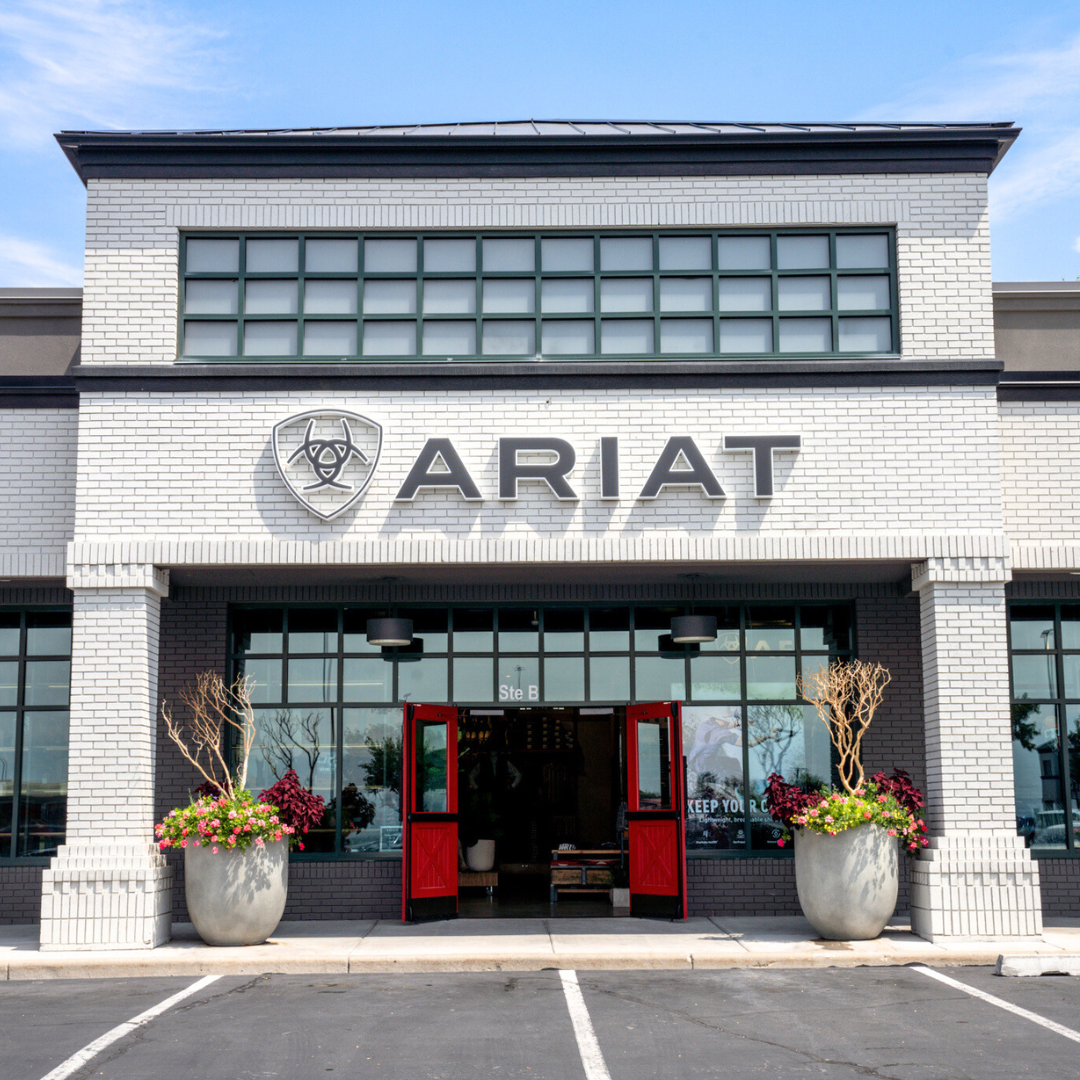 The image shows the exterior of an Ariat store. The store has a modern design with white brick and large windows. Red double doors are open, revealing the interior. The Ariat logo is prominently displayed above the entrance. The text "Shop In-Store" is overlaid on the image, serving as the hook and call to action.
