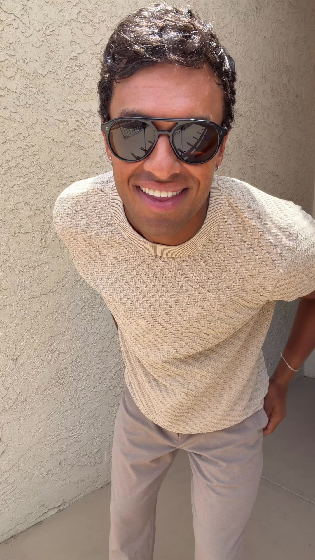 A man wearing sunglasses and a beige knit sweater with a textured pattern, and light-colored pants, smiles at the camera. He is leaning forward slightly, with his hands in his pockets. The background is a textured beige wall. Text overlays the image, with the hook "Ditch the standard shirt." and additional copy describing the product as lightweight, breathable, and luxurious.