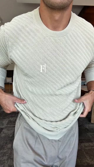 A man is shown from the chest down, wearing a light beige textured long-sleeve shirt and light grey trousers. He is pulling the hem of the shirt away from his body with both hands. A white logo with the letters "FJ" inside a rectangle is overlaid on the shirt. The text "If you only buy one long sleeve this season, make it this one. The Riviera Texture Tee Long Sleeve gives you a lightweight, breathable knit with an elevated texture that feels like luxury. Dress it down for the city or up for an event." is overlaid on the image.
