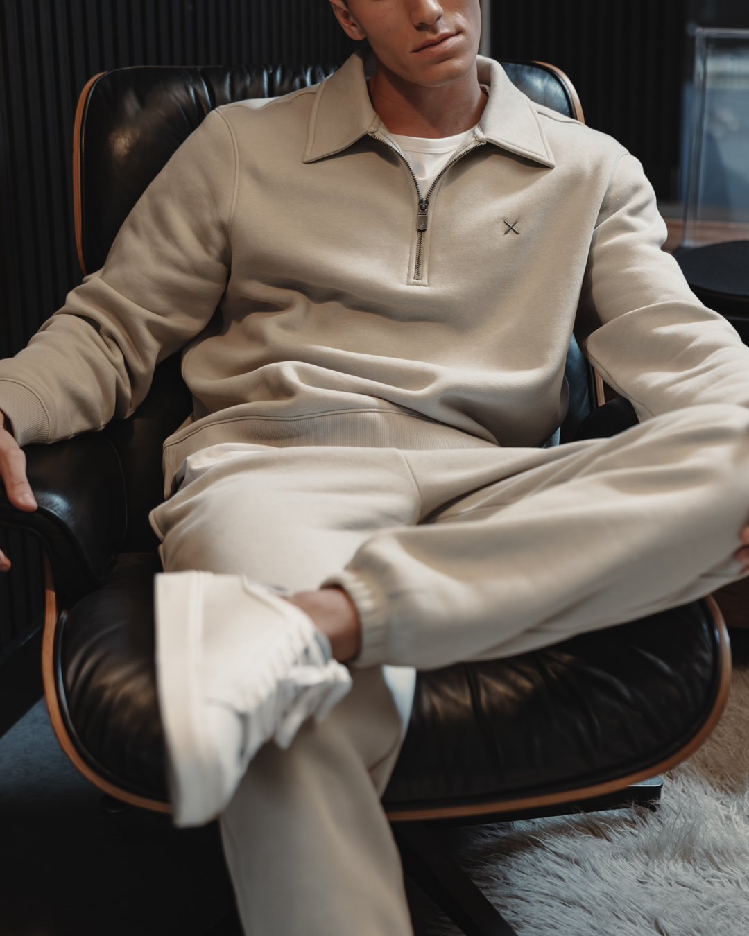A man is sitting in a black leather chair, wearing a light beige quarter-zip sweatshirt and matching sweatpants. He is looking slightly off-camera with his legs crossed, showcasing the comfortable loungewear. The text overlay announces the arrival of the 'Divine Fleece' winter collection, highlighting its warmth and softness.