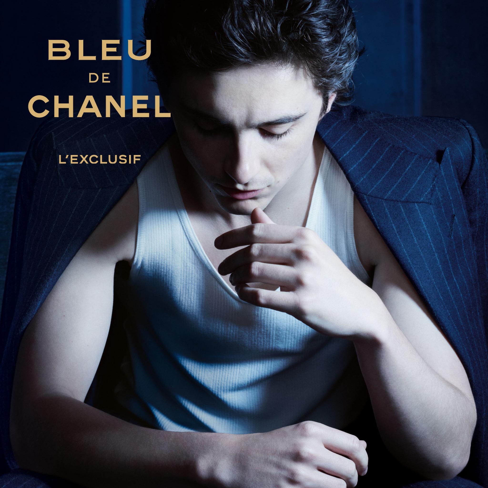 A close-up shot of a young man with dark hair, looking down with his hand near his chin. He is wearing a white ribbed tank top and a dark pinstripe suit jacket draped over his shoulders. The text "BLEU DE CHANEL L'EXCLUSIF" is displayed in gold capital letters across the top left of the image. The overall mood is sophisticated and intimate, with dramatic lighting.