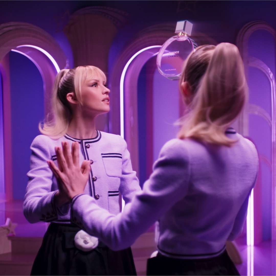The creative features a young woman with blonde hair in a ponytail, wearing a light purple jacket with black trim. She is looking towards her reflection in a mirror, which is also a woman in the same outfit. A bottle of Chanel Chance Eau Tendre perfume is suspended in the air between them. The background is a surreal, purple-lit architectural space with arched doorways. The text overlay reads "Wspaniała historia chance." and describes the film, the scent, and its availability in various sizes.