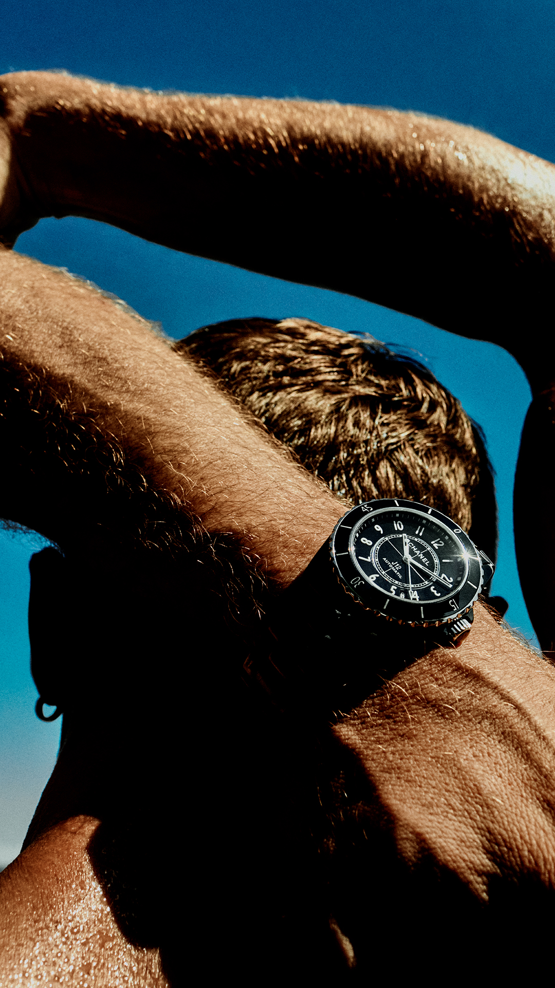 The creative shows a close-up of a man's arm and shoulder against a bright blue sky. He is wearing a black Chanel J12 watch on his wrist. The man's skin is glistening with sweat, suggesting an outdoor or athletic activity. The image focuses on the watch and the texture of the skin, with the man's head and arms partially obscured by shadow.
