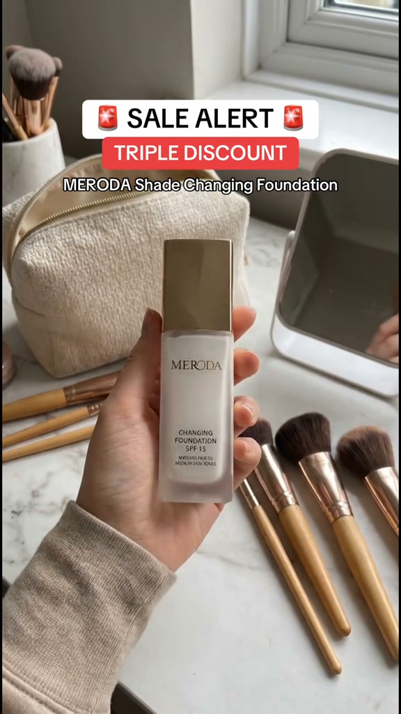 A hand holds a bottle of MERODA Shade Changing Foundation in front of a makeup bag and brushes. Text overlays read "SALE ALERT" and "TRIPLE DISCOUNT" with a siren emoji. Below that, "MERODA Shade Changing Foundation" is displayed. The foundation bottle is the main focus, with text on it indicating "CHANGING FOUNDATION SPF 15" and "NATURALLY FAIR TO MEDIUM SKIN TONES". The overall scene is set up to highlight a promotional sale on the product.