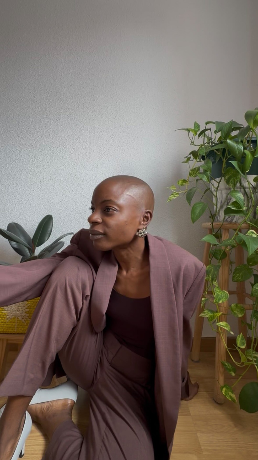 A bald woman wearing a mauve suit sits on the floor, leaning against a woven stool, with plants in the background. She looks to the left, away from the camera. The text overlay, which is part of the hook, discusses a decade change and a Caudalie product promotion, mentioning free patches and a spa treatment giveaway with a discount code. The overall aesthetic is chic and relaxed, suggesting a personal endorsement.