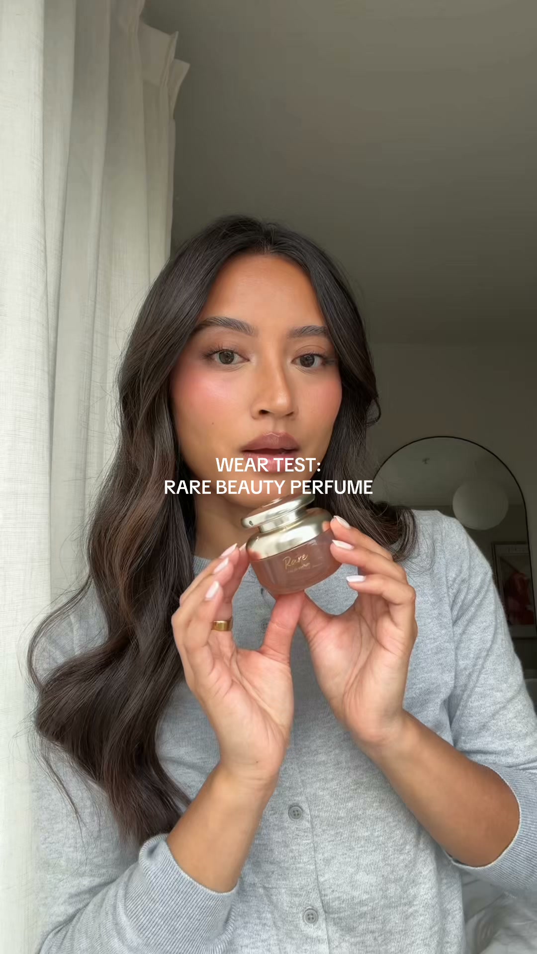 A young woman with dark hair and makeup is holding a small, ornate perfume bottle. She is looking directly at the camera with a neutral expression. Text overlaid on the image reads "WEAR TEST: RARE BEAUTY PERFUME". The background is a softly lit room with sheer curtains and a hint of a mirror. The overall impression is a personal review or demonstration of the product's longevity and scent.