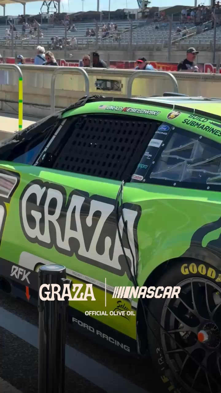 A close-up, side view of a green and black NASCAR race car is shown. The car has large white lettering that says "GRAZA" on the door, and smaller text below that reads "OFFICIAL OLIVE OIL". The "GRAZA" logo is also visible on the side of the car, along with the NASCAR logo. The tires have "GOOD" written on them. The background shows a blurred view of a racetrack with spectators in the stands.
