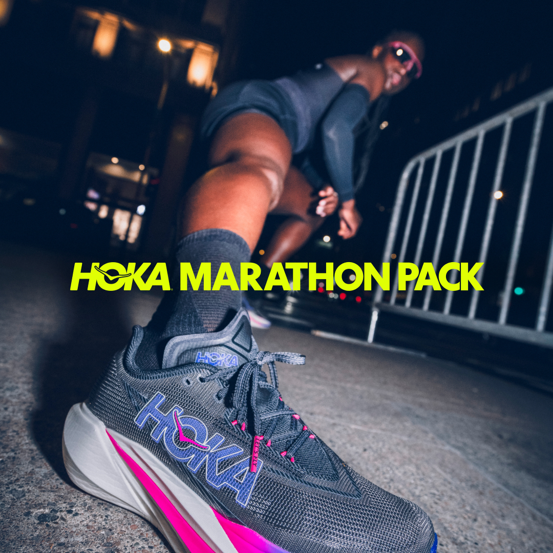 A low-angle, dynamic shot focuses on a Hoka running shoe in the foreground, with a runner blurred in the background, appearing to be in motion. The shoe is grey with vibrant pink and blue accents. Bright yellow text overlaid on the image reads "HOKA MARATHON PACK." The scene is set at night, with streetlights and a metal barrier visible, suggesting an urban running environment.