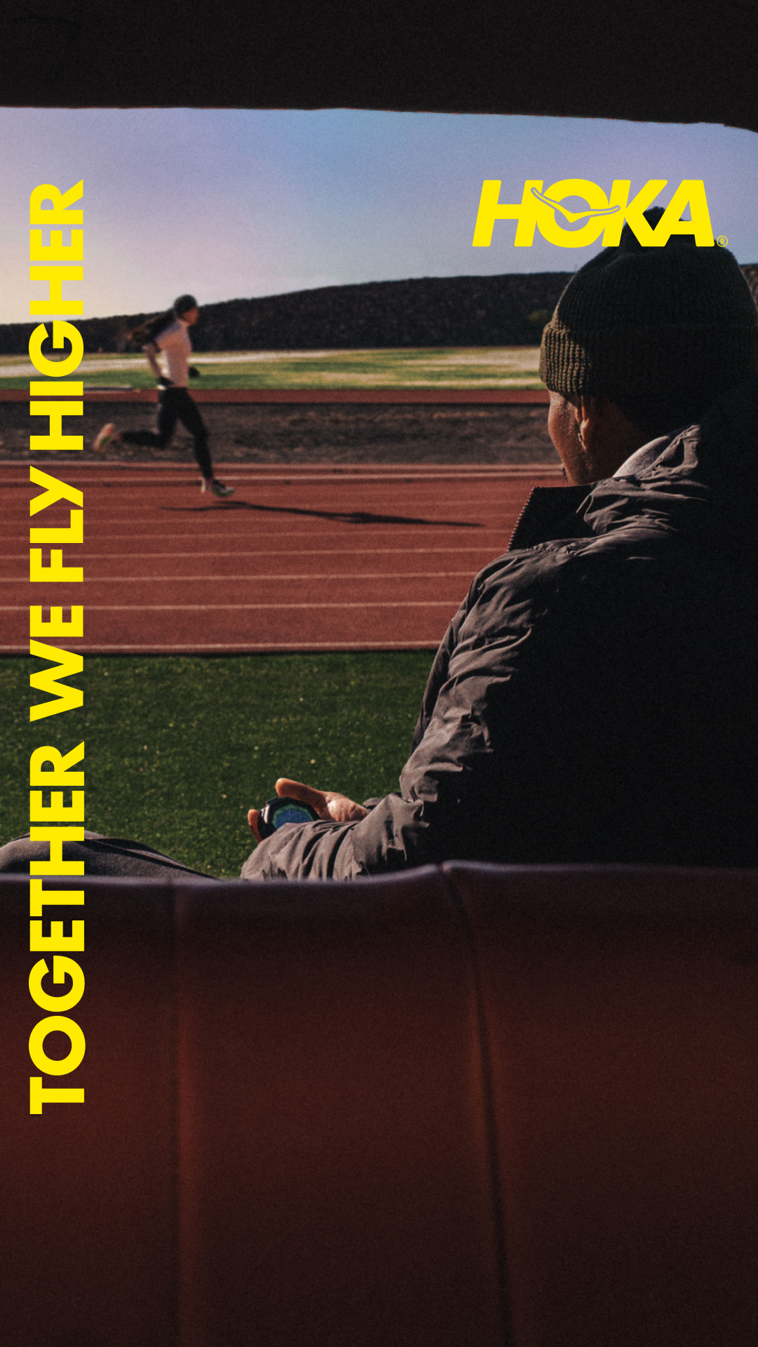 The creative shows a person sitting in what appears to be a stadium or bleachers, looking out at a running track. In the background, a runner is in motion on the track. The Hoka logo is visible in the top right corner. Bold yellow text on the left side of the image reads "TOGETHER WE FLY HIGHER." The overall scene evokes a sense of shared ambition and athletic pursuit.
