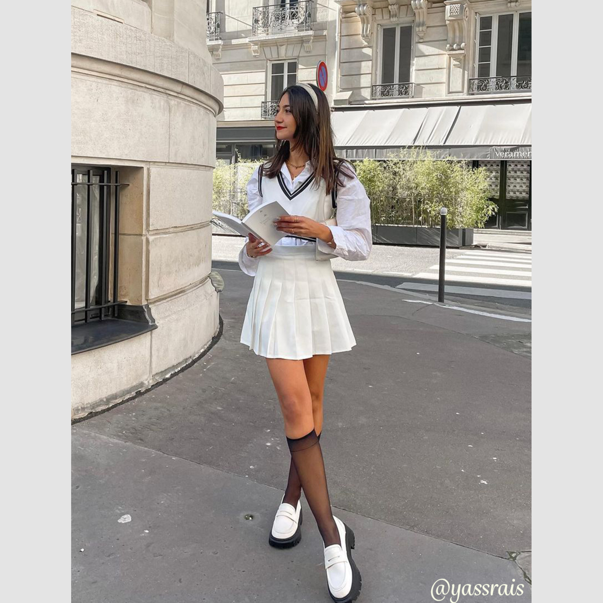 A young woman stands on a Parisian street, holding a book and looking to the side. She is wearing a white collared shirt, a white V-neck sweater vest, and a white pleated mini-skirt, paired with black sheer knee-high socks and white loafers. The text "too niche? we don't think so 😉snag something special today! Enjoy 20% off EVERYTHING for new users on the Cider app🎉 ✈️ We ship globally" is overlaid on the image.