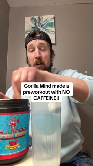 A man wearing a black baseball cap is shown from the chest up, mixing a blue liquid in a clear bottle with a scoop. To his left is a large blue and red tub of Gorilla Mind pre-workout powder. White text overlaid on the image reads: "Gorilla Mind made a preworkout with NO CAFFEINE!!" The man appears to be speaking directly to the camera, though his mouth is partially obscured by his hand.