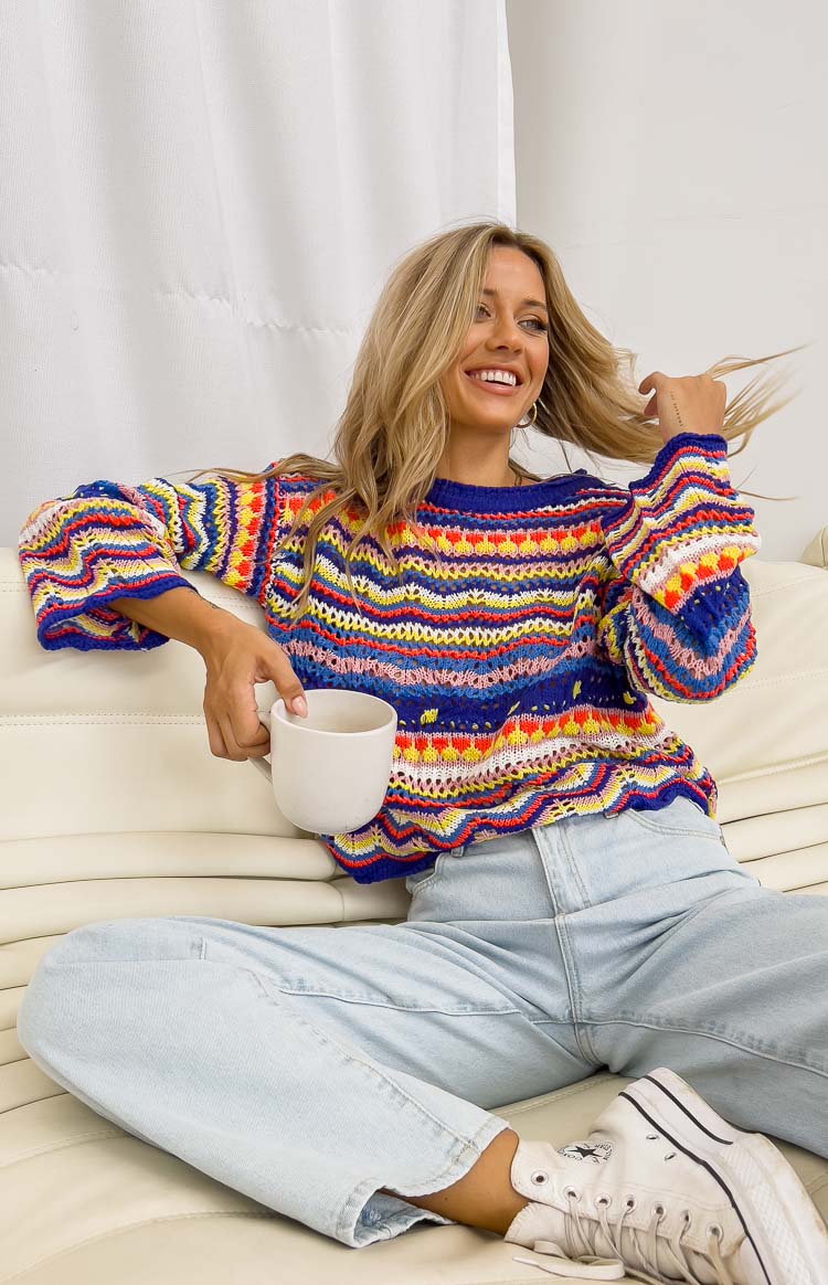 A smiling woman with blonde hair sits on a white couch, holding a white mug. She is wearing a colorful, striped, crocheted sweater and light blue jeans. Text overlays the image, with the main hook "Don't let your new fave outfit pass you by!" prominently displayed. The ad encourages viewers to shop best-sellers online.