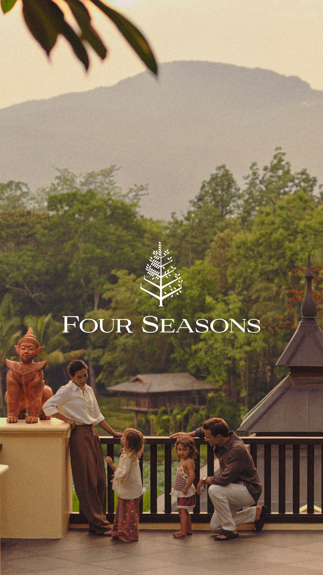 A serene, golden-hour scene unfolds on a balcony overlooking lush, green mountains and trees. A family, including two young children and their parents, are gathered, with the parents interacting with the children. The Four Seasons logo and name are prominently displayed in white text across the middle of the image. The overall impression is one of peaceful family enjoyment in a beautiful, natural setting.