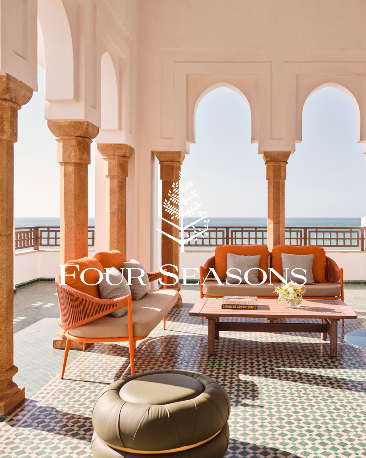 A luxurious outdoor patio with a view of the ocean. The patio features modern orange outdoor furniture, including a sofa, chairs, and a coffee table. The text "FOUR SEASONS" is overlaid in large, elegant white font across the center of the image. The scene evokes a sense of upscale relaxation and comfort, suitable for a high-end resort or hotel.